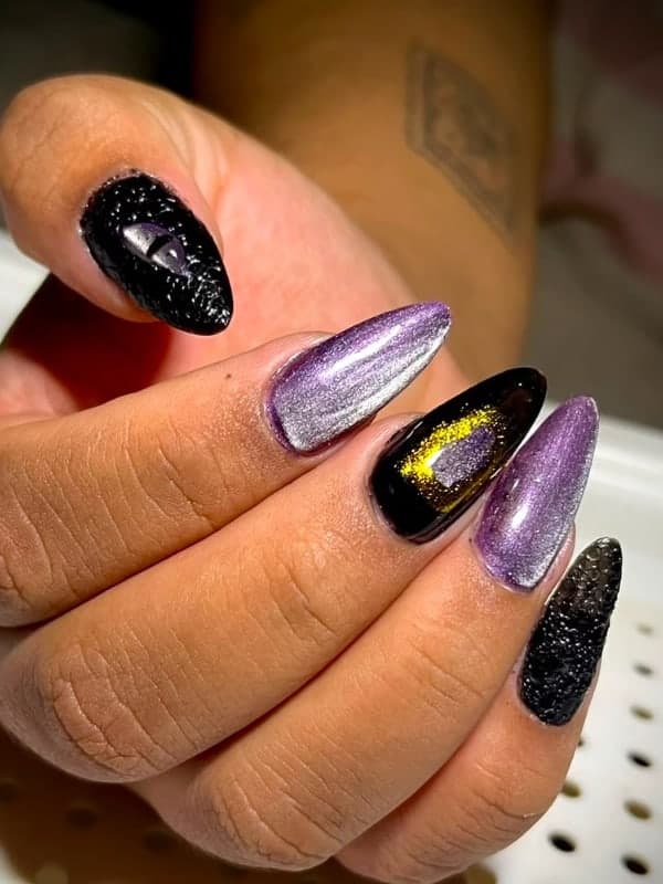 Mão fechada com unhas amêndoa em gel magnético lilás perolado, uma unha preta com risco dourado e outra preta texturizada